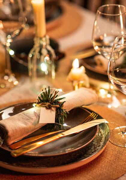 Weihnachtlich gedeckter Tisch mit einer Serviette, Messer, Gabel, Teller, Unterteller, Weingläsern und brennenden Kerzen.