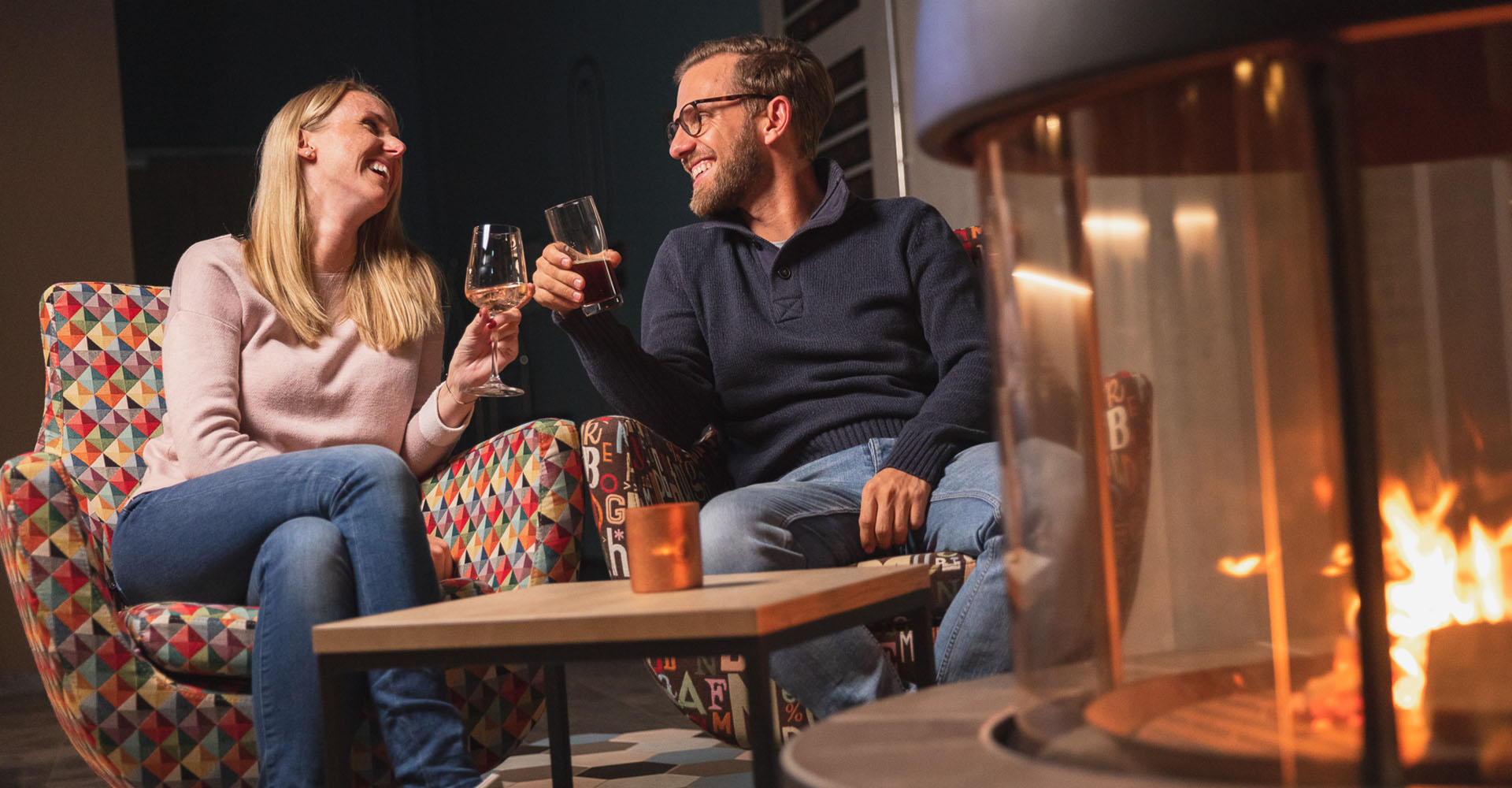 Ein Pärchen sitzt an einem modernen Kamin und stößt mit einem Glas mit Getränk an. Die blonde Frau lacht, der Mann mit der Brille lächelt sie an.