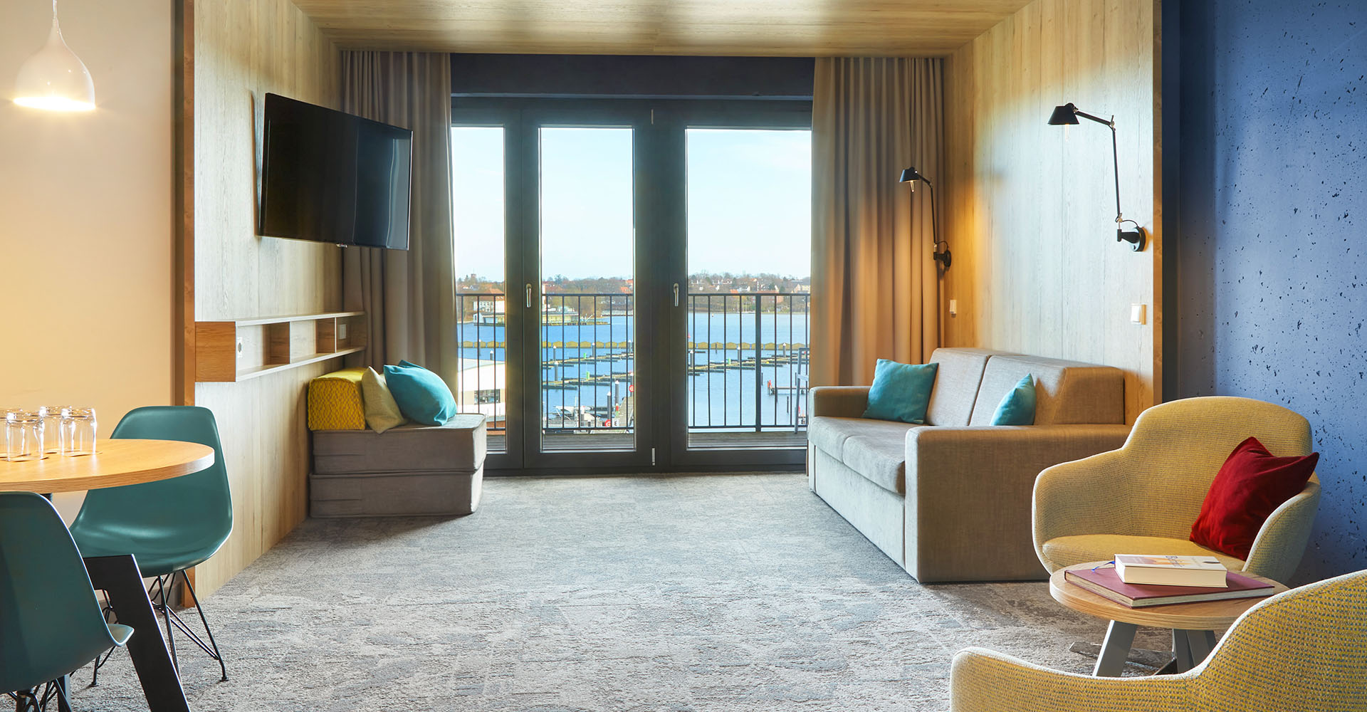 Der Wohnbereich in der ancora Suite im arborea Marina Resort Neustadt mit Sofa, Sesseln, TV, kleinem Essbereich mit Tisch und Stühlen sowie Balkon mit Blick auf die Ostsee.