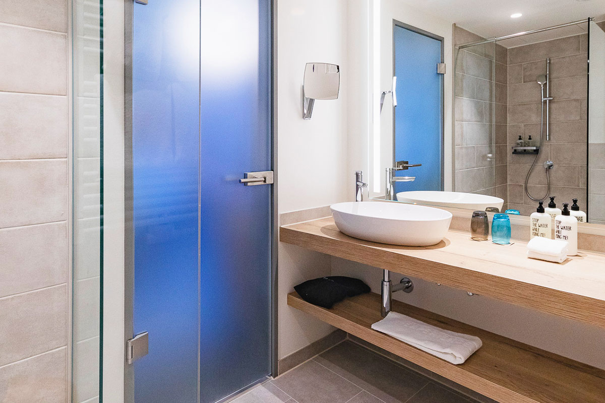 Badezimmer mit Kosmetikspiegel, Dusche und Waschbecken inklusive Körperpflegeprodukte von RITUALS und Handtücher im arborea Marina Resort Neustadt.