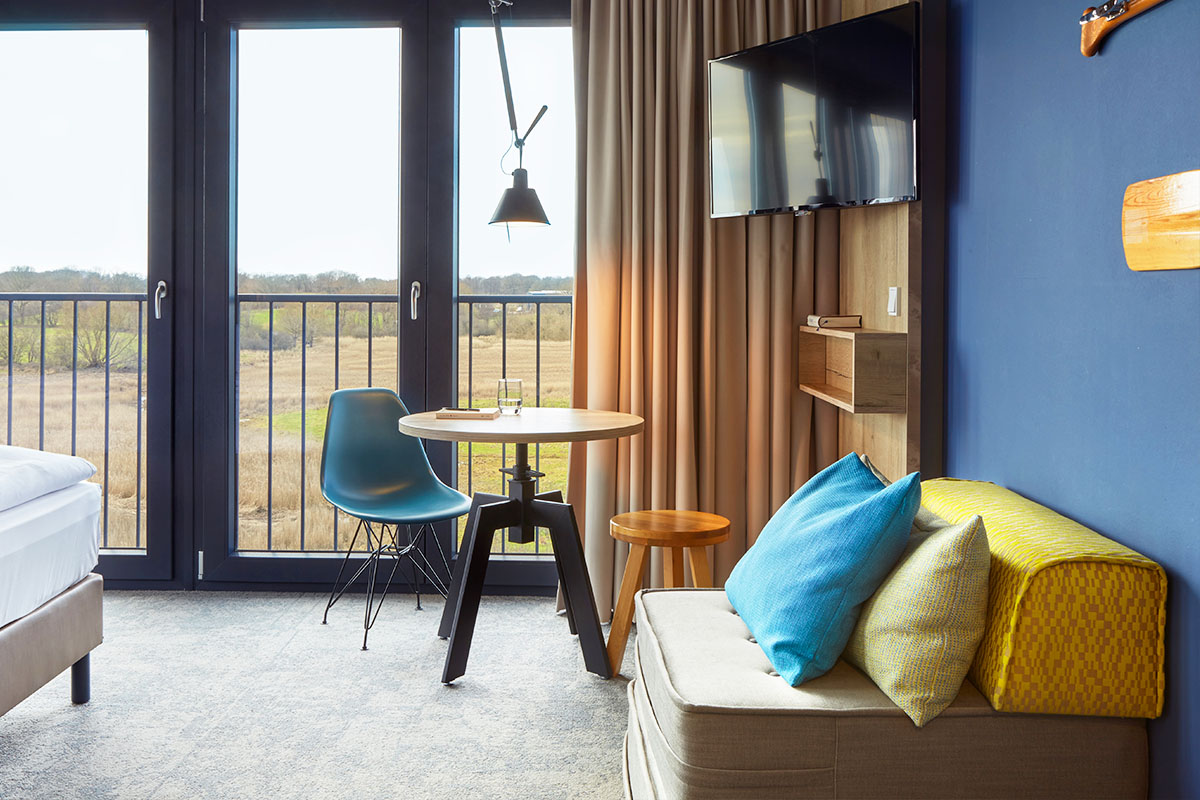 Gemütlicher Wohnbereich mit einem Sessel, einem kleinen Esstisch mit Stühlen und einem Ausblick auf die Landseite im Doppelzimmer Landblick im arborea Marina Resort Neustadt.