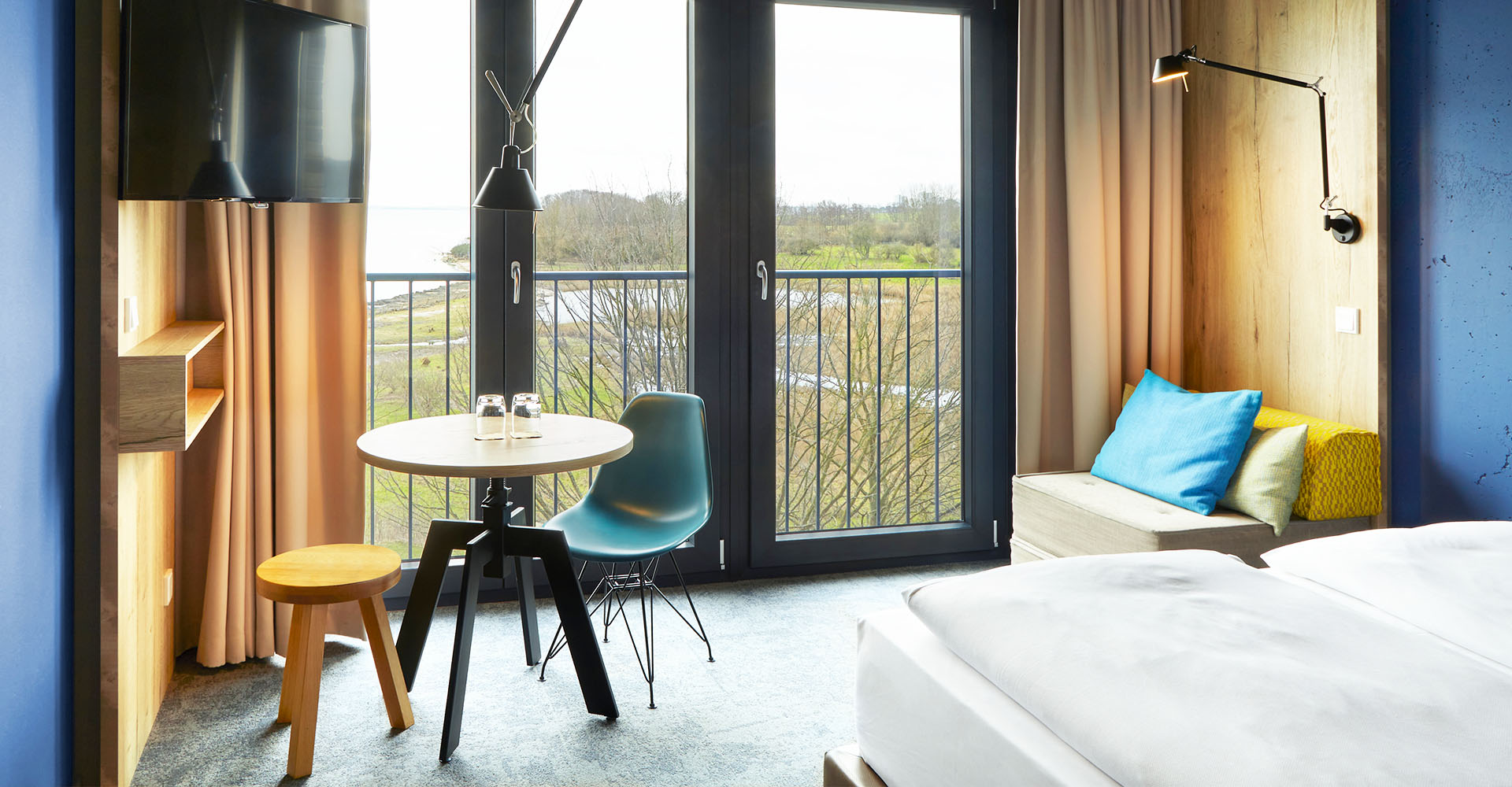 Doppelzimmer im arborea Marina Resort Neustadt , mit einem gemütlichen Kingsize Doppelbett, einem Sessel, einem kleinen Esstisch mit Stuhl und Hocker, einem TV und großen Fenstern mit Landblick.